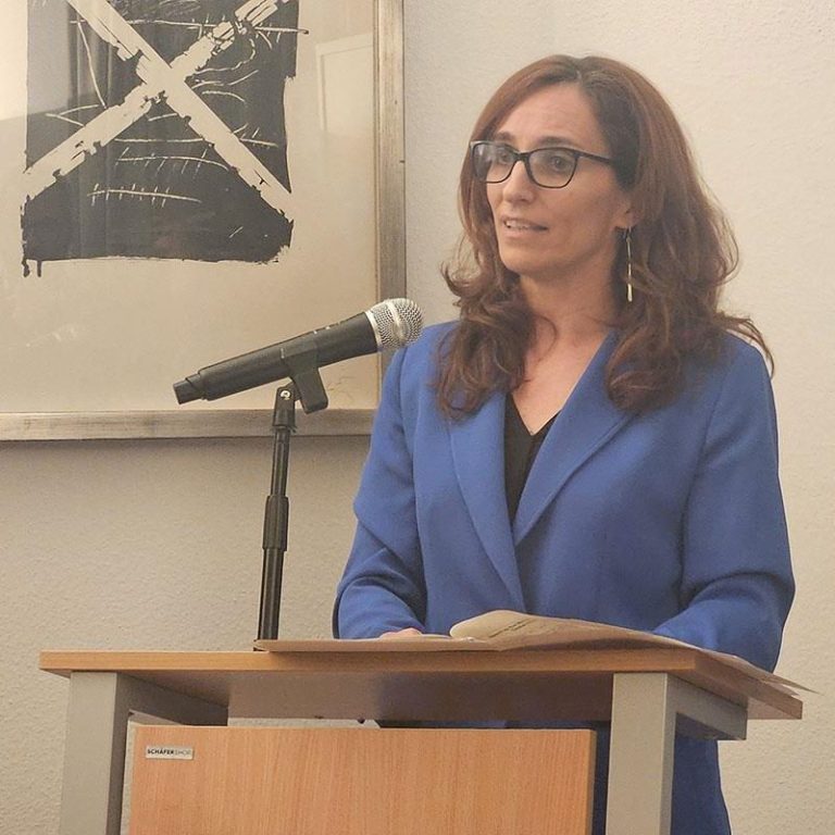 Mujer con chaqueta azul, sonriente, ante un micrófono en un podio.